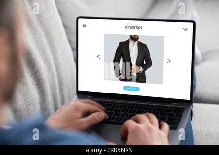Young man choosing new suit on laptop screen, enjoy online shopping remotely from home, sitting on sofa Stock Photo