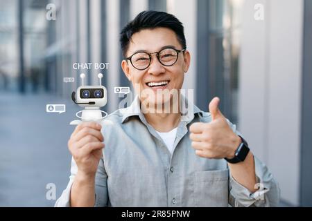 Cheerful middle aged asian man holding Chatbot virtual icon, collage Stock Photo