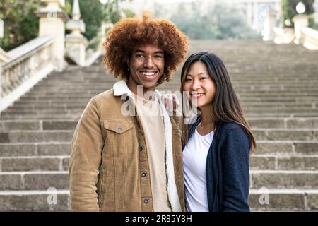 Lovely multiracial couple looking at camera in a park.Two happy diverse young persons smiling and looking at camera outside. Stock Photo