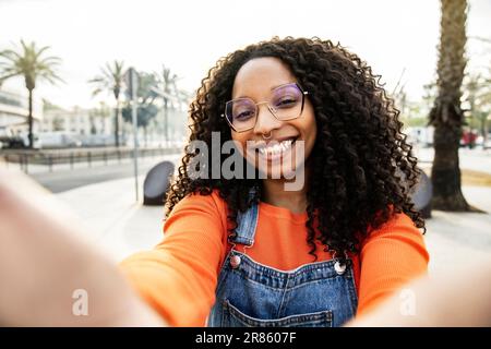 Portrait of beautiful happy young woman smiling wearing glasses and a piercing looking at camera taking a selfie in the street. happy young curly hair Stock Photo