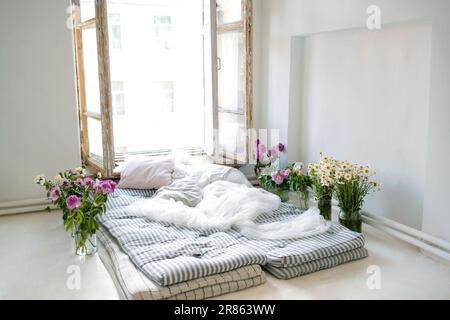 Minimalistic retro bedroom interior. Mattresses, pillows and a bedspread on the floor near the huge window. Vases with peonies and daisies along the s Stock Photo