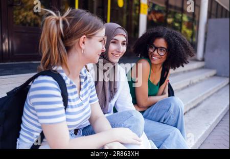 Multiracial female students laugh and chat on the college or university steps outfoors. Happy friends with backpacks are talking and having fun. Cauca Stock Photo