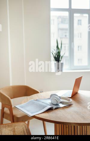 A cozy freelancer workplace on a round wooden table in a minimalistic bright interior of the house. Headphones, magazine and laptop on the table. A po Stock Photo