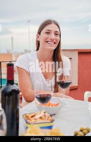 Vertical portrait of a joyful young woman celebrating a lunch with wine and some snacks, looking at camera sitting at table on a rooftop terrace at house. Smiling beautiful lady enjoying a nice day. High quality photo Stock Photo