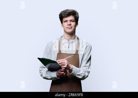 Young guy in apron making notes on clipboard, on white background Stock Photo