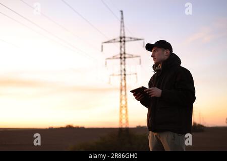 Young power engineer holds digital tablet against high voltage tower at field. Electrical engineer checking energy efficiency electrical towers. World Stock Photo