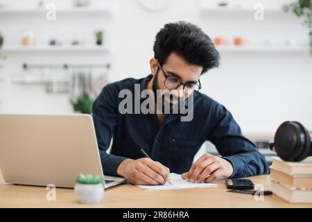 Focus on man's left hand with pen making notes on draft of paper document being placed near laptop on wooden desk. Well-organised arabian remote employee starting business day in workplace at home. Stock Photo