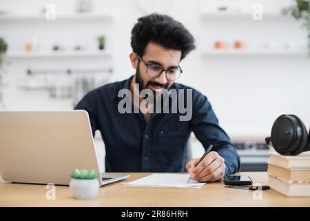 Focus on man's left hand with pen making notes on draft of paper document being placed near laptop on wooden desk. Well-organised arabian remote employee starting business day in workplace at home. Stock Photo