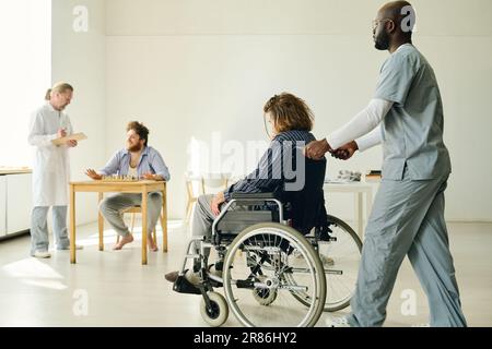 Young African American man in uniform pushing wheelchair with mentally ill guy while moving along ward where doctor talking to male patient Stock Photo