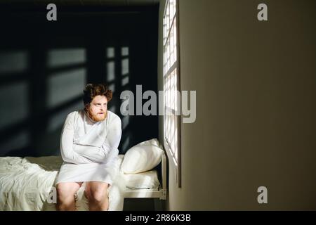 Young insane male patient of mental hospital in straitjacket sitting on bed and looking through window with sunlight penetrating into ward Stock Photo