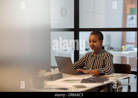 Young serious African American female designer typing on laptop keyboard while sitting by workplace in office and working over new project Stock Photo