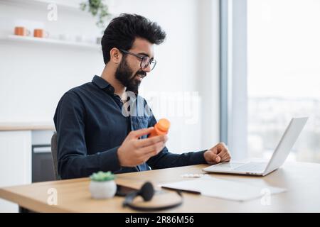 Handsome mature person in spectacles holding bottle of pills while sitting in home office with laptop at midday. Indian businessman examining medicine prescribed after online consultation by doctor. Stock Photo