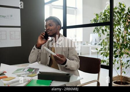 Young confident black man in casualwear talking to colleague on mobile phone while sitting by desk with sketches, catalog and office supplies Stock Photo