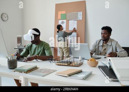 Young African American webdesigner looking at laptop screen while sitting by workplace next to colleague in headset creating new computer games Stock Photo