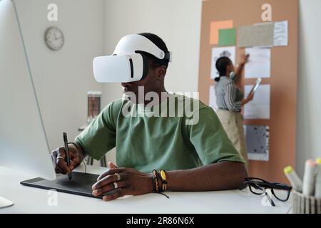 Young African American male designer with vr headset using touchpad and stylus while sitting by desk and creating new computer games Stock Photo