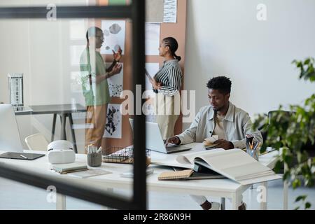 Young serious male designer of interior with color swatches in hand sitting by workplace in front of laptop and creating new graphic drawings Stock Photo