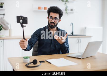 Focus on modern cell phone placed in tripod being held by tired indian man in glasses sitting at desk with laptop. Professional consultant live streaming detailed report from distant workplace. Stock Photo