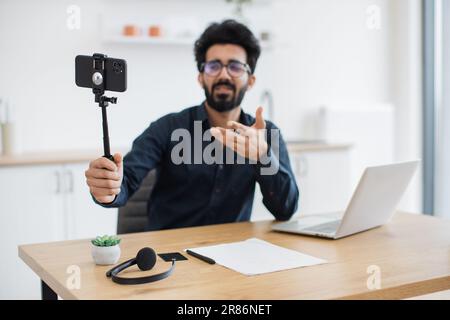 Focus on modern cell phone placed in tripod being held by tired indian man in glasses sitting at desk with laptop. Professional consultant live streaming detailed report from distant workplace. Stock Photo