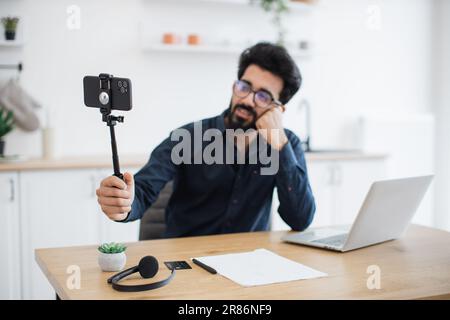Focus on modern cell phone placed in tripod being held by tired indian man in glasses sitting at desk with laptop. Professional consultant live streaming detailed report from distant workplace. Stock Photo
