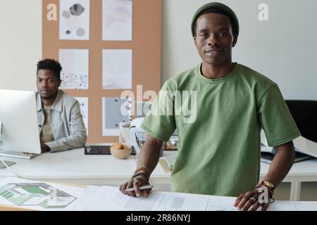 Young confident male architect in casualwear standing by workplace with blueprints and looking at camera during work over new project Stock Photo