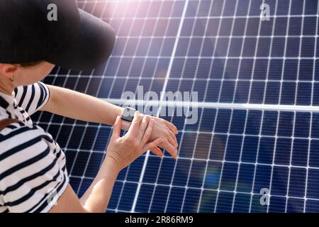 Woman in cap using her smartwatch on her hand on background with solar energy panel. Stock Photo