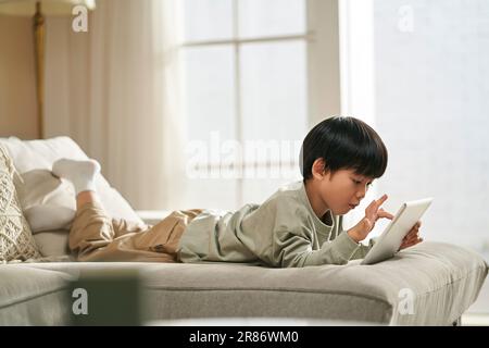 five-year-old little asian boy lying on front on family couch playing computer game using digital tablet Stock Photo