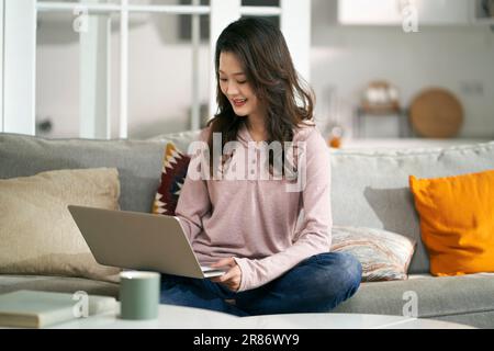 https://l450v.alamy.com/450v/2r86wy9/young-asian-professional-woman-sitting-on-couch-at-home-working-with-laptop-computer-2r86wy9.jpg