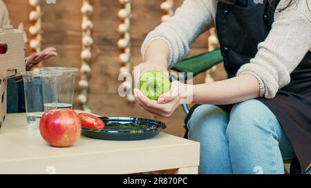 Small business owner cutting apples to give sample to cheerful clients at food festival, sitting at table. Woman stand holder doing product sampling at farmers market. Handheld shot. Close up. Stock Photo