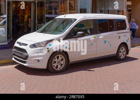 White Ford Transit minivan, with decals of bees and flowers, parallel parked in front of a store on the Santa Fe Plaza, Santa Fe, New Mexico, USA. Stock Photo