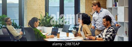 Office life. Group of young multiracial people sitting at the table in coworking space and working together, using modern technologies and discussing Stock Photo