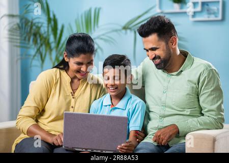 Indian couple got happy by seeing son using laptop while sitting together at home - concept of generation, family time and parenthood. Stock Photo