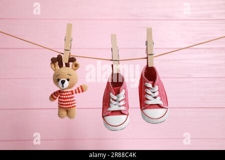 Cute baby sneakers and crochet toy drying on washing line against pink wooden wall Stock Photo