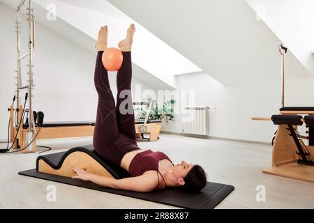 A young girl is engaged in Pilates in a bright studio. A slender brunette in a burgundy bodysuit is doing exercises on a yoga mat with a reformer and Stock Photo