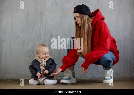 Young long haired mom in red hoodie and black baseball cap plays with her cute baby in beige hoodie and funny motorcycle jacket sitting on the floor. Stock Photo