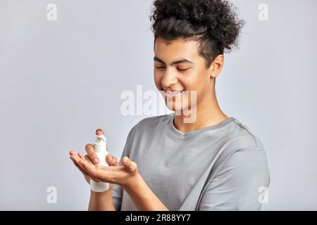 Young handsome smiling guy with black curly hair squeezing skincare cream from the dispenser to his hand. Biracial man in studio with healthy skin pro Stock Photo