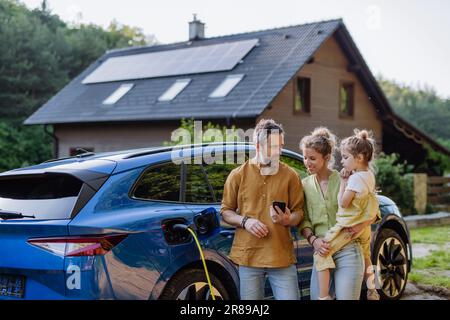 Family with little girl standing in front of their house with solar panels on the roof, having electric car. Stock Photo