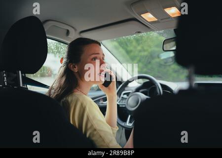 Scared young woman sitting in electric car with drained battery, making a phone call to the service center. Stock Photo