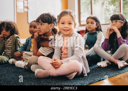 Adorable little child looking at the camera while sitting in class with other kids. Girl attending a lesson in elementary school. Primary education an Stock Photo