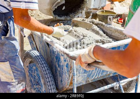 Teamwork, workers are pouring fresh mortar in wheelbarrow from mortar mixer machine. Construction site works Stock Photo