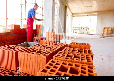 Close up shot on heap of red blocks, in background mason is building a wall with red ceramic blocks using mortar from a plastic trough. Building is un Stock Photo