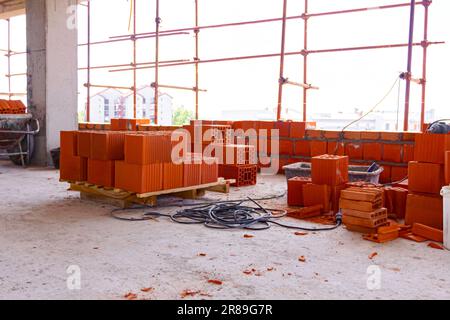 Wooden pallet with pile of red blocks, in the background is wall installed with red ceramic bricks and mortar, building is under construction. Stock Photo
