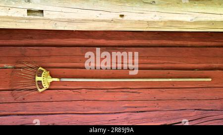 Old rusted rake hanging on the exterior of traditional log wall painted with red ochre paint, just below eaves. Kreivinmaki Open-Air Museum, Salo, FI. Stock Photo