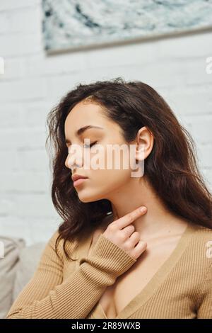 Young brunette woman in casual clothes closing eye while doing self-massage of thyroid gland on neck and promoting lymph flow at home, self-care ritua Stock Photo