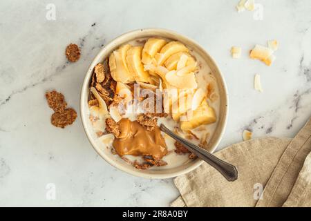 Whole grain flakes with banana, coconut chips and peanut butter in a bowl, white background, top view. Healthy breakfast concept. Stock Photo