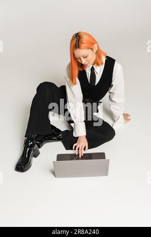 high angle view of trendy asian woman in white shirt, black vest, tie and pants sitting and networking on laptop on grey background, business casual f Stock Photo