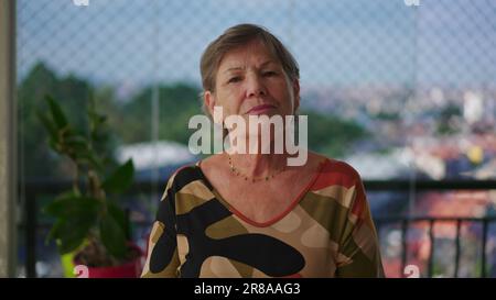 Portrait of a senior woman looking at camera with serious expression standing at apartment balcony with city view in background Stock Photo