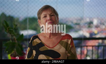 Portrait of a senior woman looking at camera with serious expression standing at apartment balcony with city view in background Stock Photo