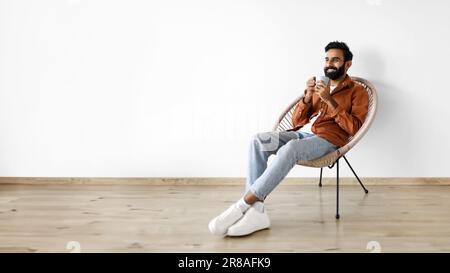 Cheerful indian man drinking coffee while chilling in armchair indoor Stock Photo