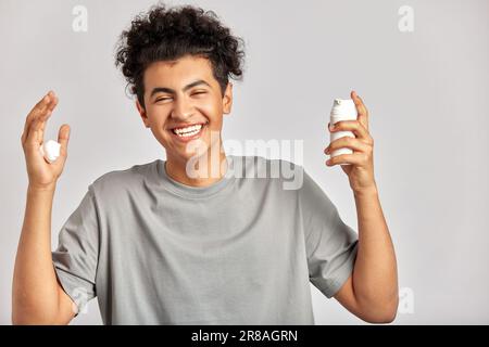 Young handsome smiling guy with black curly hair squeezing skincare cream from the dispenser to his hand. Biracial man in studio with healthy skin pro Stock Photo