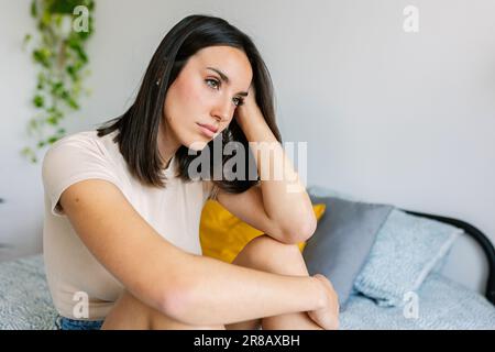 Portrait of young adult woman feeling sad sitting on bed feeling sad at home Stock Photo